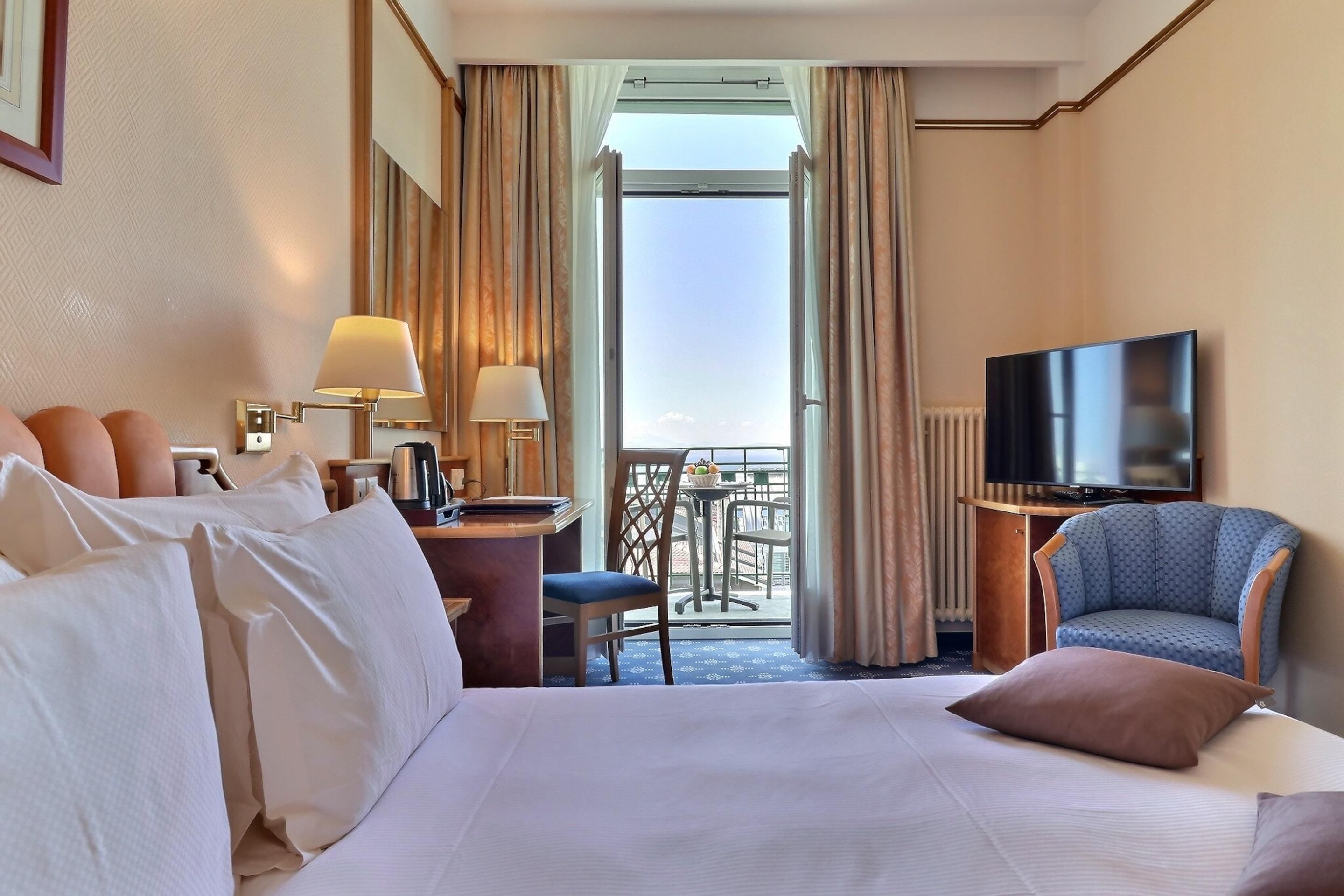 A hotel room with a bed, a desk with a lamp, a television, and a balcony with a table and chairs.