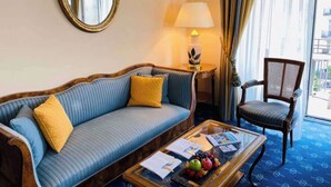 A hotel room with a blue sofa, a wooden armchair, a glass coffee table with magazines and fruit, a lamp, and a large mirror on the wall. - Hotel Mirabeau, BW Signature Collection (Lausanne)