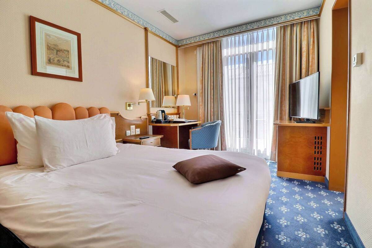 Une chambre d’hôtel avec un grand lit, un bureau avec une chaise, une télévision et une fenêtre avec des rideaux.
