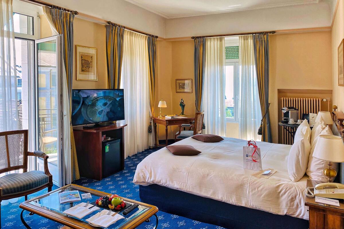 Une chambre d’hôtel avec un grand lit, un bureau avec une chaise, une télévision, une machine à café et une fenêtre avec des rideaux.