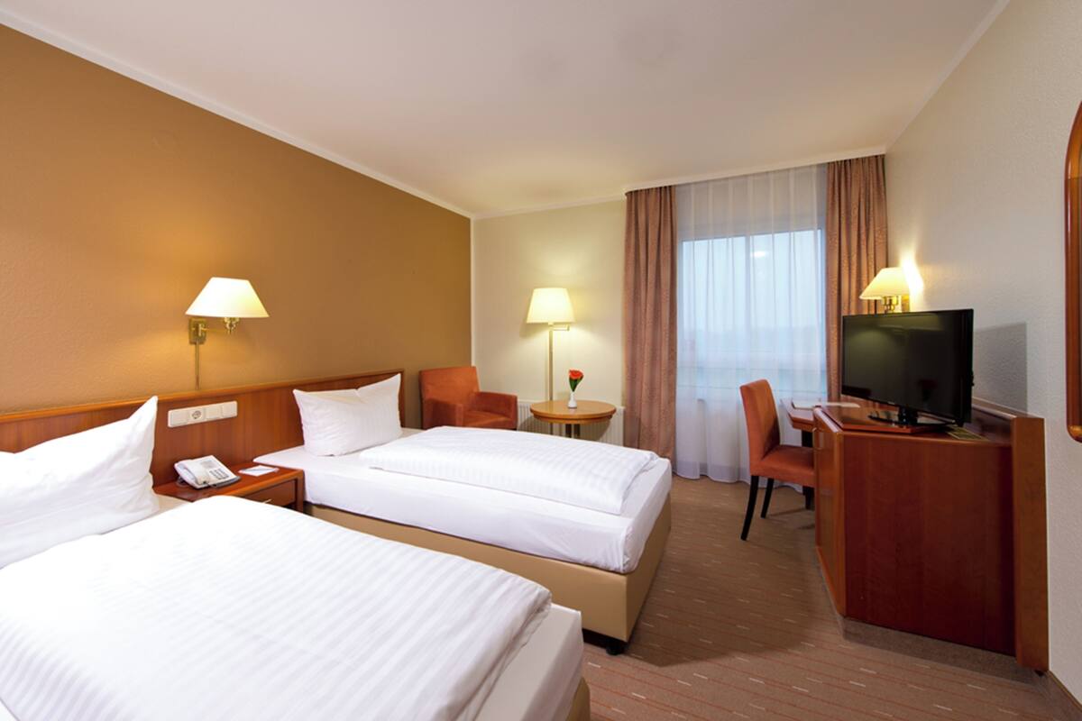 Une chambre d’hôtel avec deux lits, un bureau avec une télévision, une chaise, une petite table avec une lampe et une fenêtre avec des rideaux.