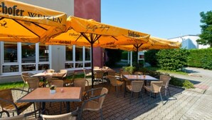 An outdoor seating area with tables and chairs under yellow umbrellas, a building with large windows, and a paved walkway. - ACHAT Hotel Schwarzheide Lausitz (Schwarzheide)