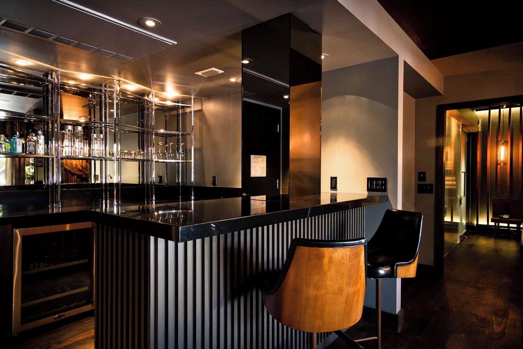 A modern bar area with a black countertop, bar stools, and a shelving unit displaying bottles and glasses.