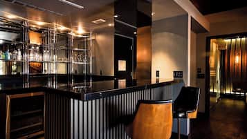 A modern bar area with a black countertop, bar stools, and a shelving unit displaying bottles and glasses.