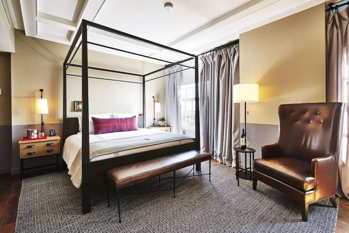 A four-poster bed with a bench, a leather armchair, a bedside table with a lamp, and a nightstand with a lamp.
