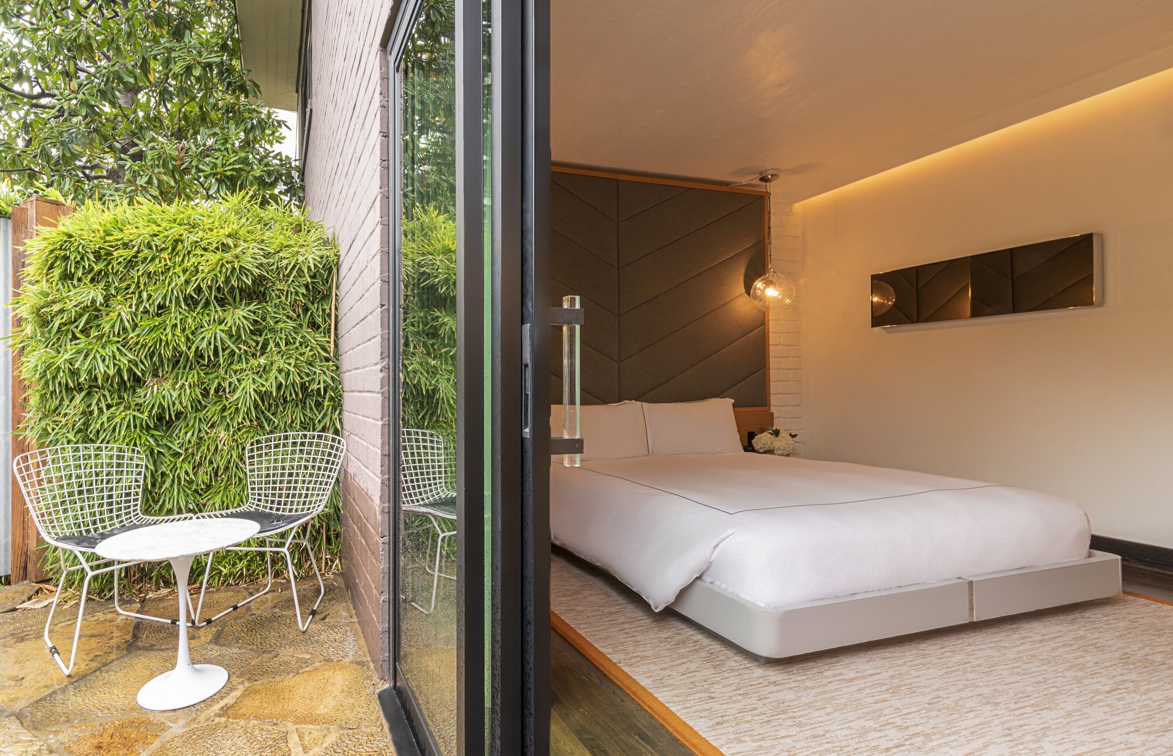 A modern bedroom with a large bed, a glass door leading to a patio with chairs and greenery.
