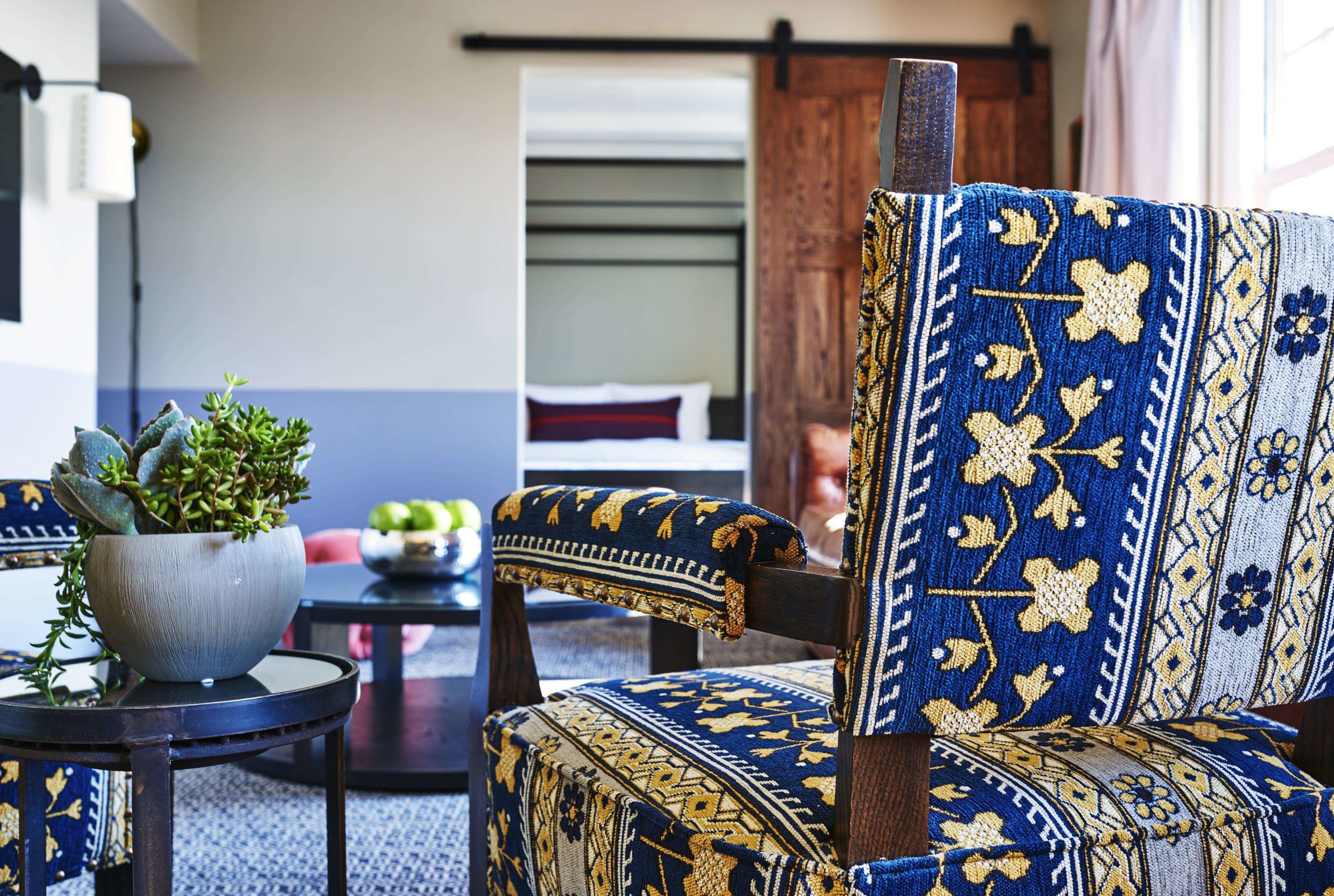 A room with a patterned armchair, a small table with a potted plant, and a bed with a red pillow in the background.