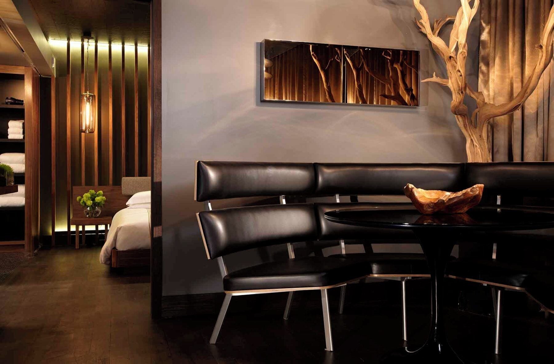 A modern hotel room with a dark wood floor, a black leather sofa, a round table, and a wooden sculpture.
