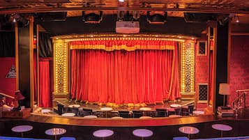A theater stage with a red curtain, a central lighted column, and a seating area with round tables.