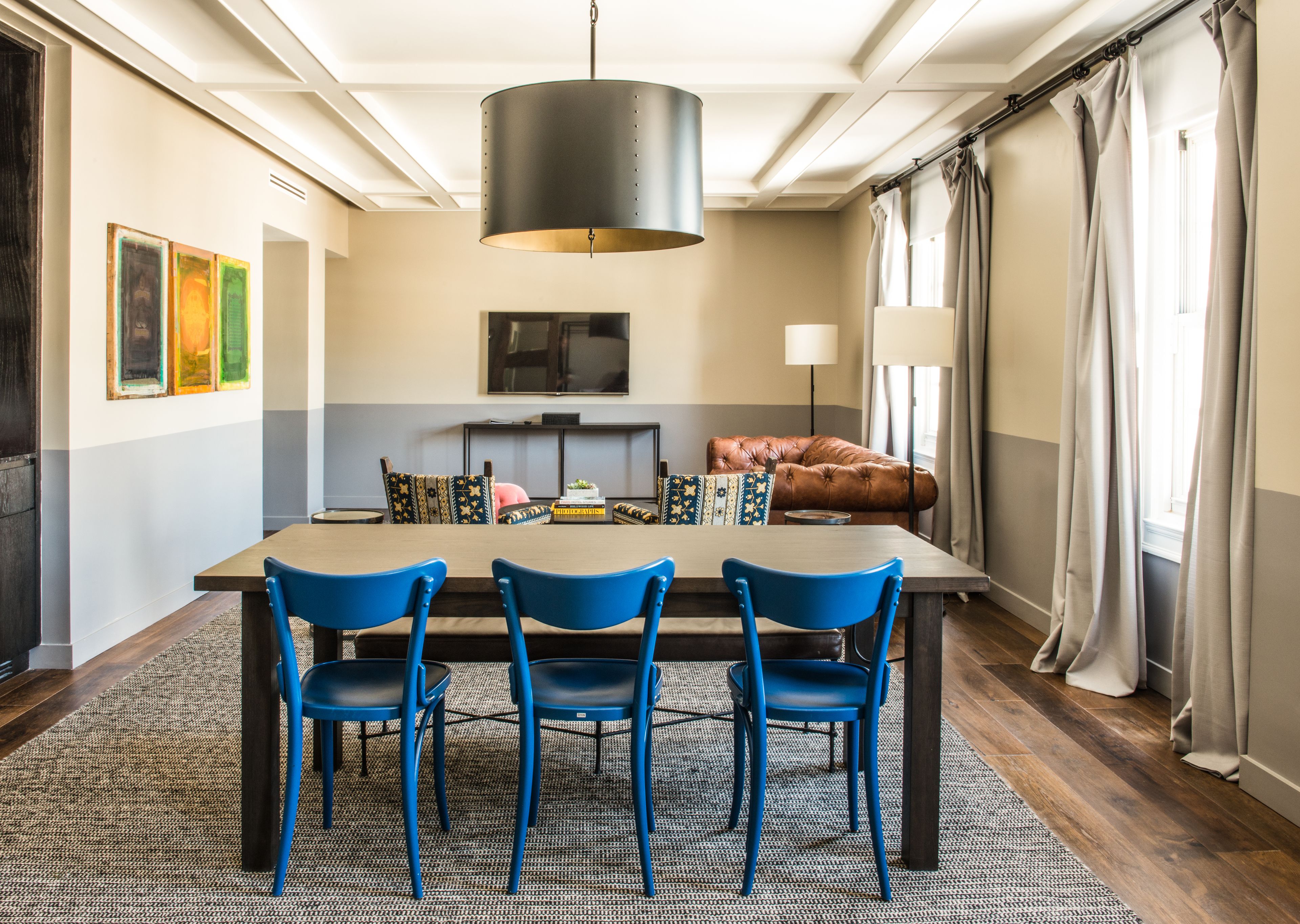 A dining area with a wooden table, three blue chairs, a black pendant light, and a sofa with patterned cushions.