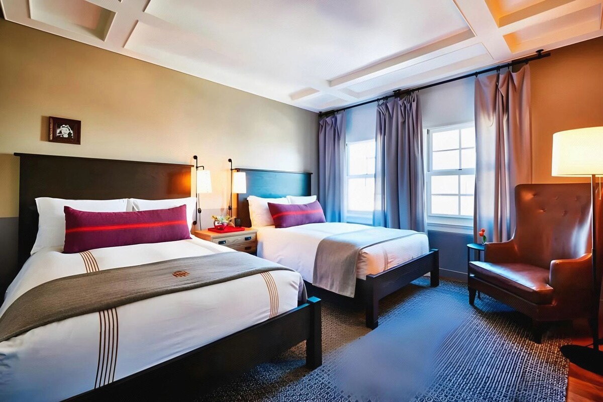 A hotel room with two beds, a chair, a nightstand, a lamp, and a window with curtains.