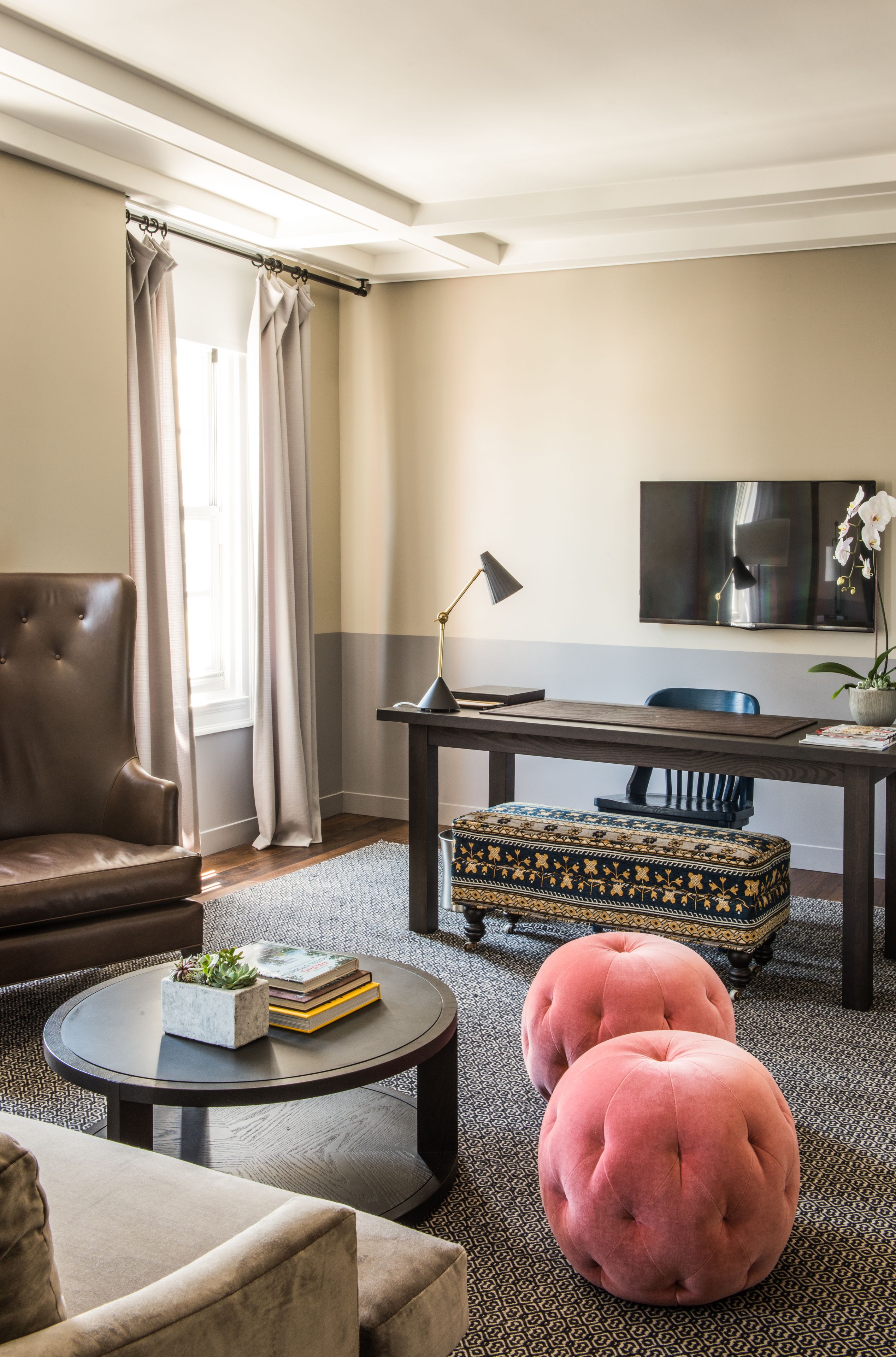Una sala de estar con un sillón de cuero, una mesa redonda de centro con libros y una planta, un reposapiés a juego, un escritorio de madera oscura, una lámpara negra y un televisor en la pared.
