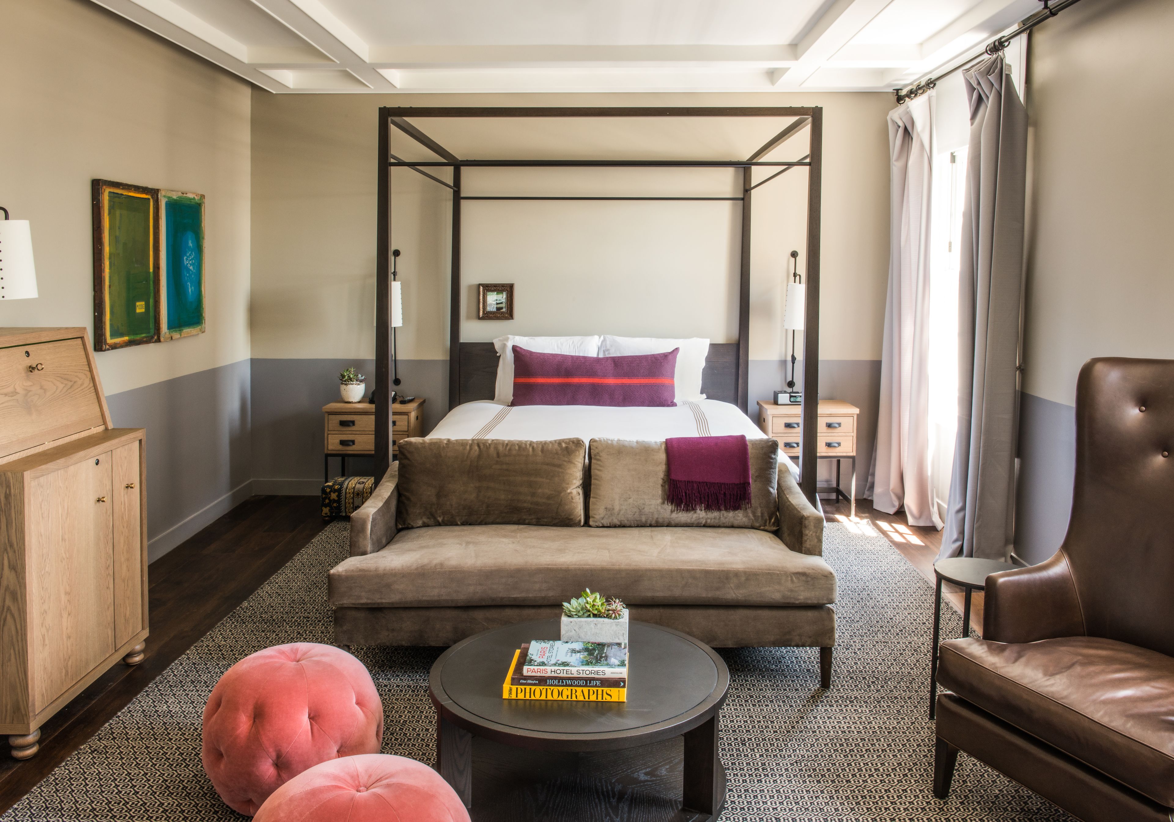 Una cama con dosel, decoración en tonos neutros, un sofá, una mesa de centro con libros y un sillón de cuero en una habitación luminosa con alfombra estampada.