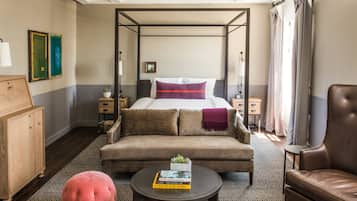 Una cama con dosel, decoración en tonos neutros, un sofá, una mesa de centro con libros y un sillón de cuero en una habitación luminosa con alfombra estampada.
