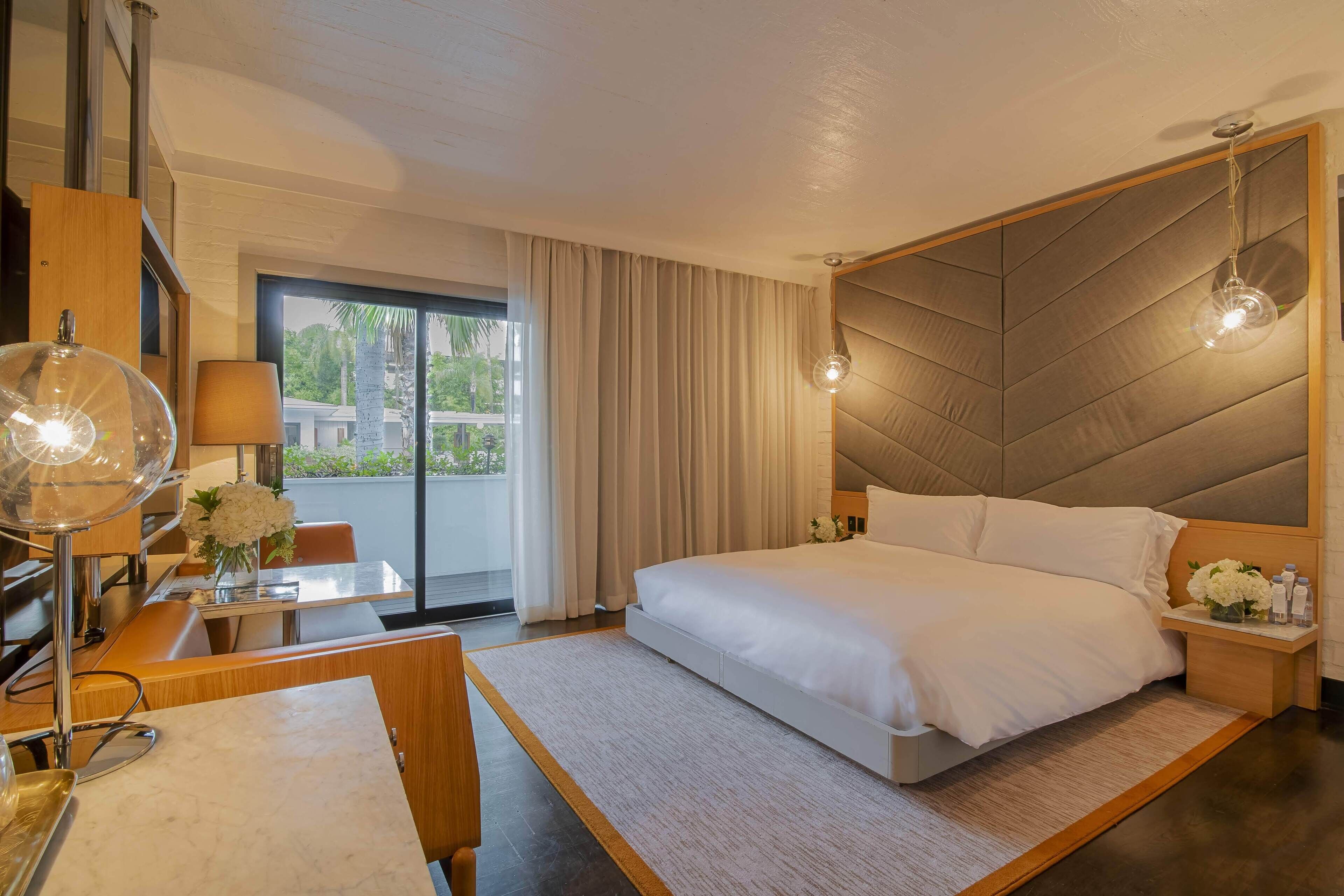A modern hotel room with a large bed, a desk with a lamp, a view of the outdoors, and a large window with curtains.