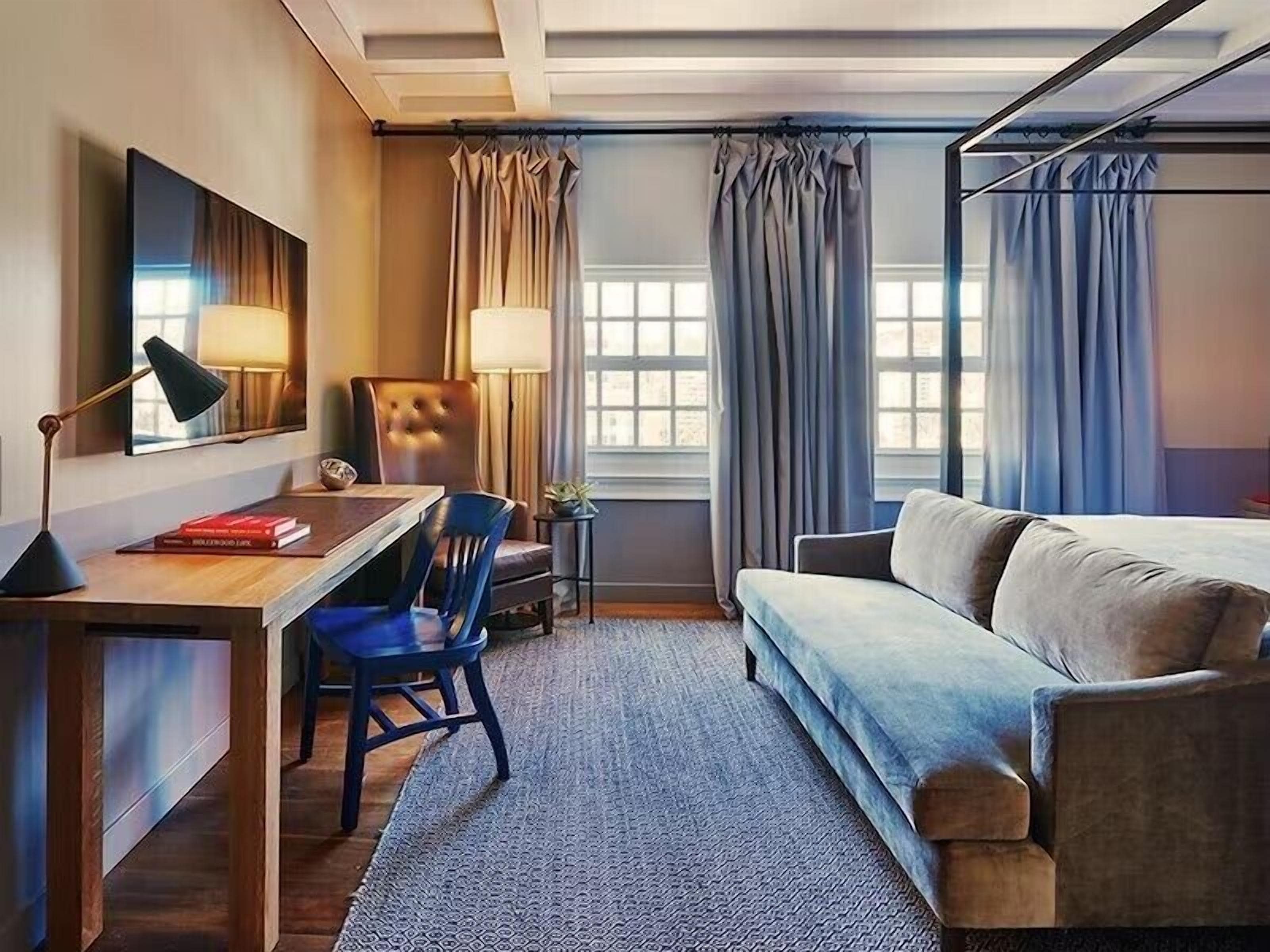 Een hotelkamer met een bureau, stoel en bank. Er is een raam met gordijnen, een lamp en een spiegel aan de muur.
