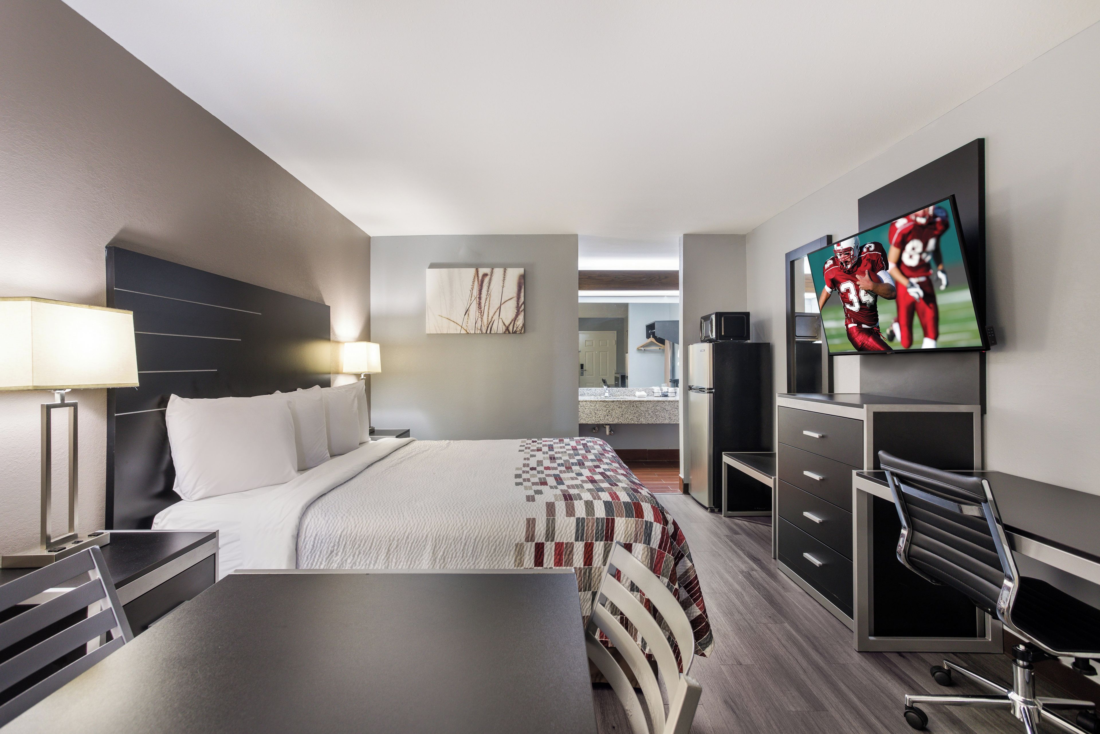 A modern hotel room with a bed, bedside table, lamp, flat-screen TV, desk, chair, and a view of the bathroom and kitchen.
