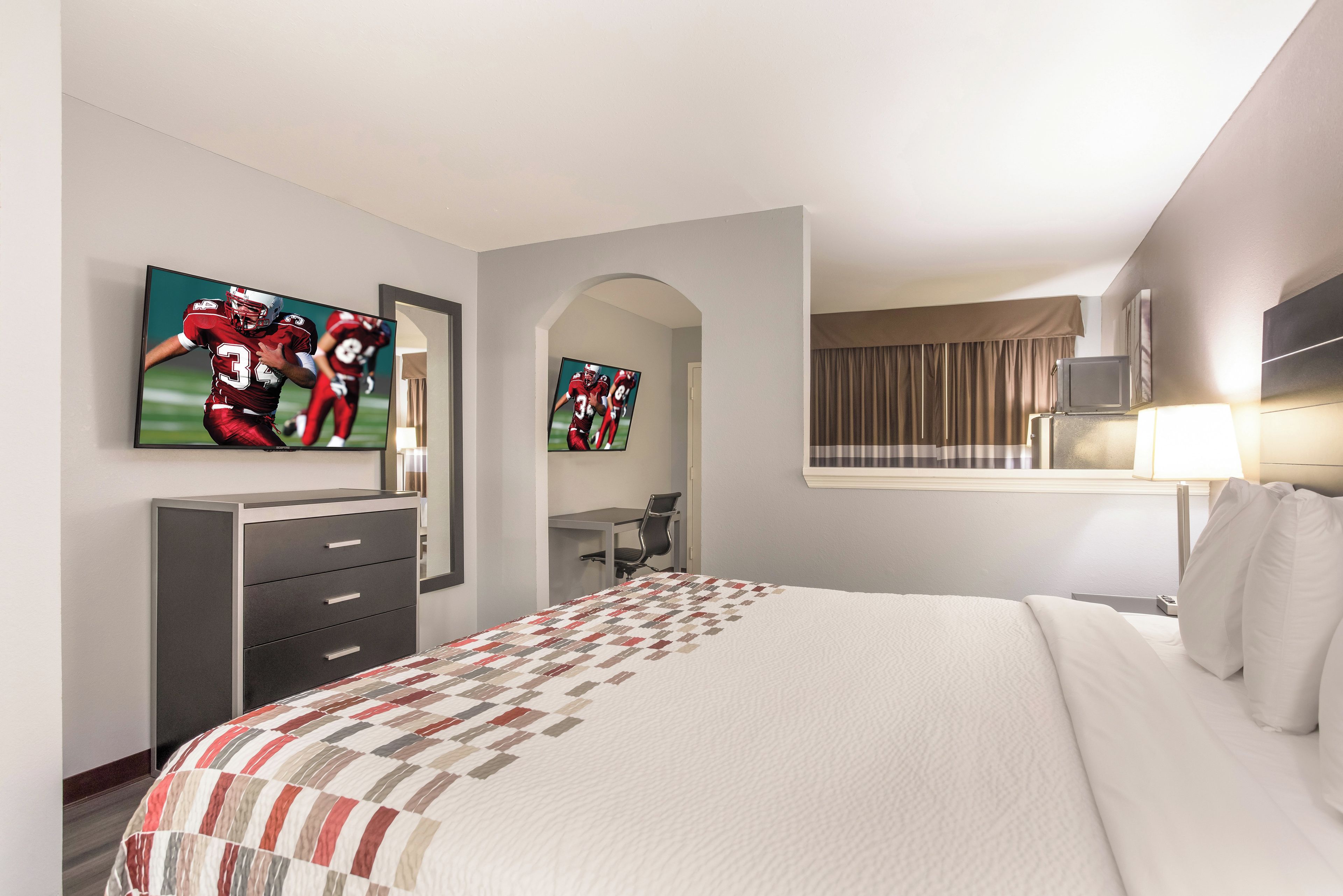 A hotel room with a large bed, a desk, a chair, a television mounted on the wall, and a window with curtains.