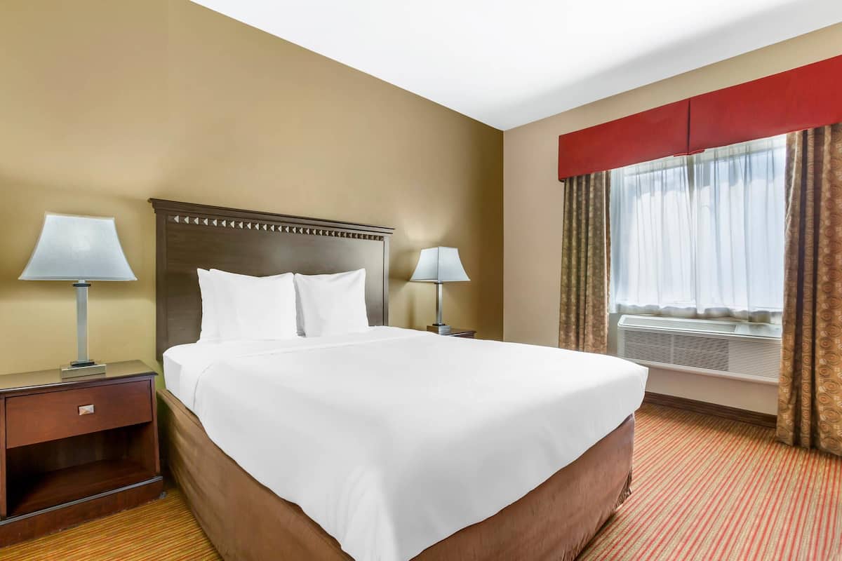 A hotel room with a large bed, two bedside tables with lamps, a window with curtains, and an air conditioning unit.
