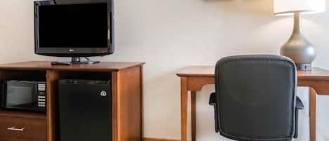 A hotel room with a wooden desk, a black office chair, a television, a mini-fridge, a lamp, and two framed pictures on the wall.