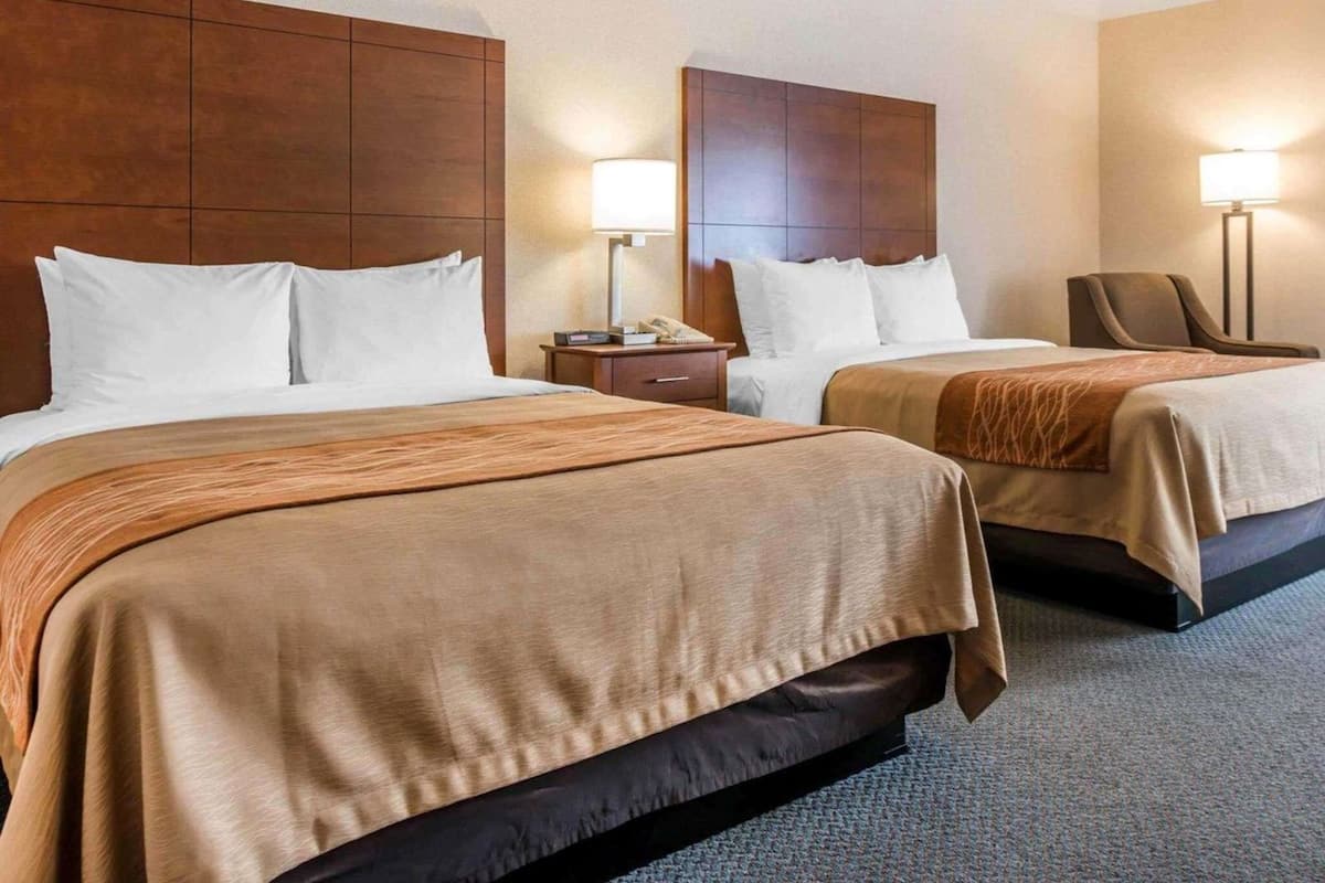 A hotel room with two beds, wooden headboards, bedside lamps, a desk, and a chair.