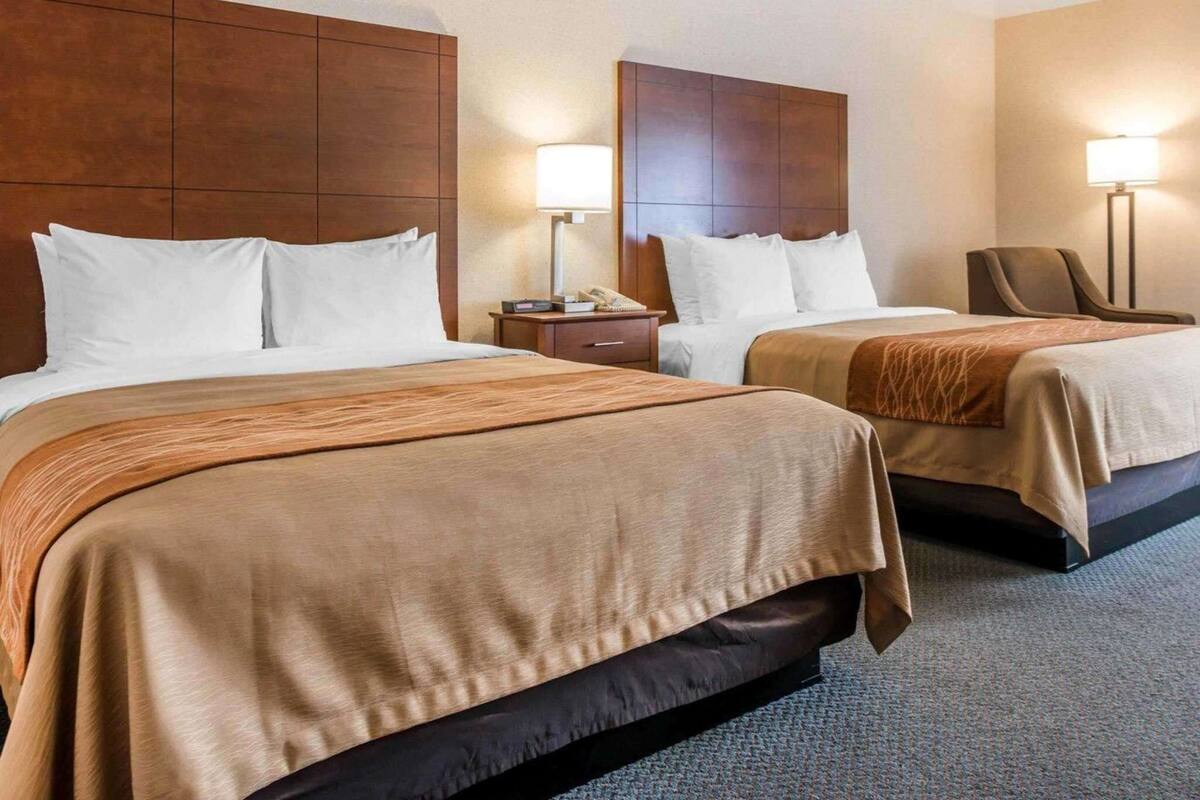 A hotel room with two beds, wooden headboards, bedside tables with lamps, and a chair.