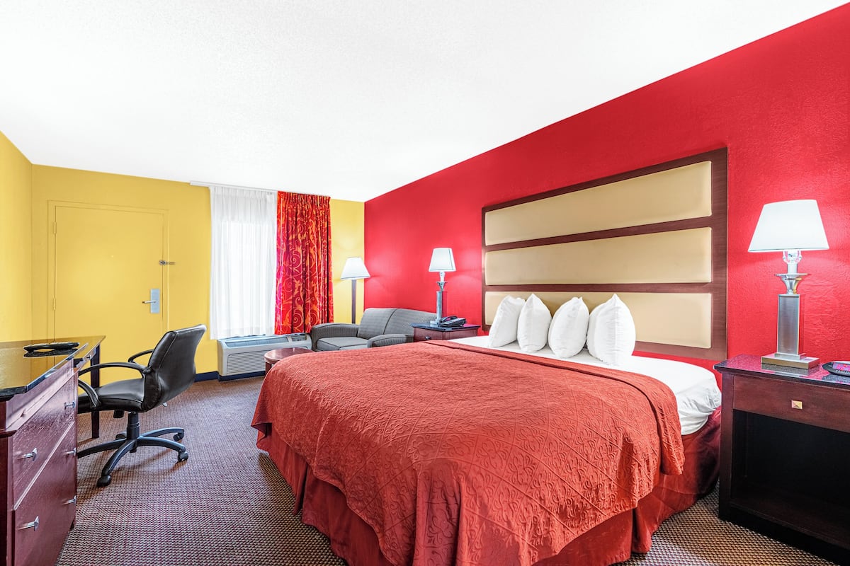Quarto de hotel com esquema de cores vermelho e amarelo, uma cama grande com almofadas brancas, uma escrivaninha com cadeira e duas luminárias de cabeceira.