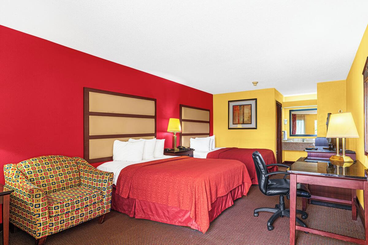 Quarto de hotel com esquema de cores vermelho e amarelo, uma cama, uma escrivaninha com cadeira, uma mesinha de cabeceira com abajur e uma poltrona estampada.