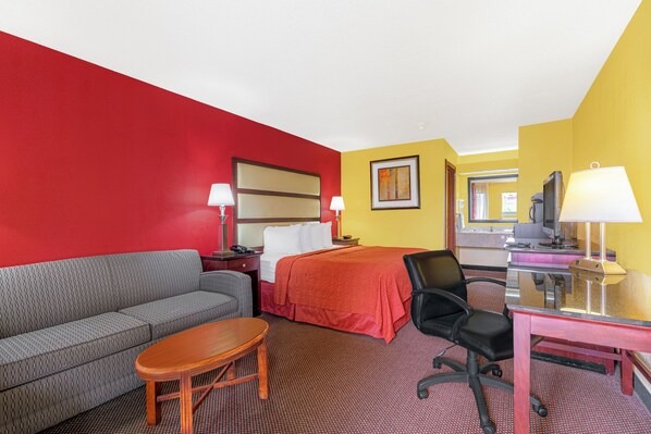 A hotel room with a red wall, a gray sofa, a wooden table, a bed with a red bedspread, a black office chair, and a desk with a lamp. - Welcome Inn (Milledgeville)