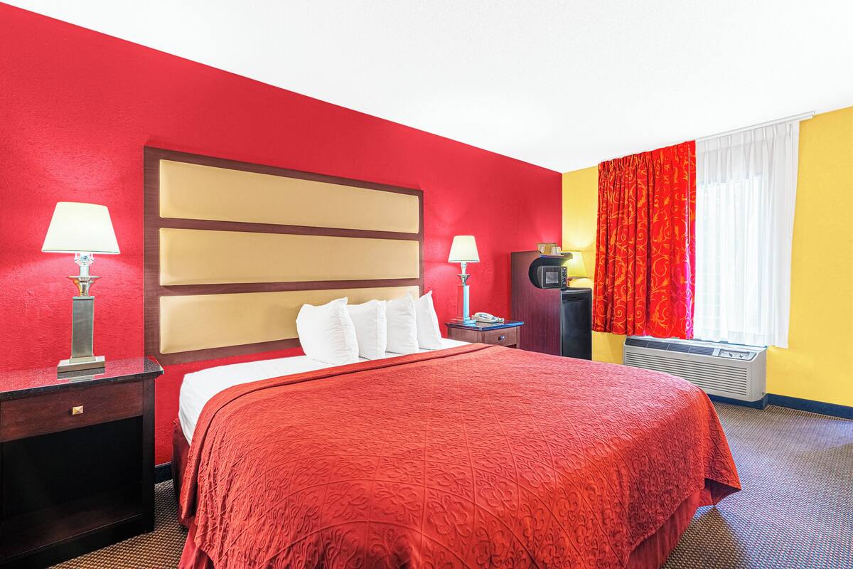 Quarto de hotel com esquema de cores vermelho e amarelo, uma cama grande com roupa de cama branca, duas mesas de cabeceira com abajures, uma televisão e uma janela com cortinas vermelhas.