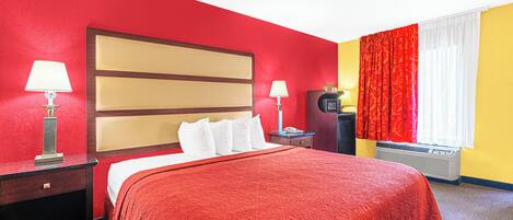 Habitación de hotel con una decoración en tonos rojo y amarillo, una cama con almohadas blancas, una mesita de noche con lámpara, un escritorio con teléfono y una ventana con cortinas rojas.