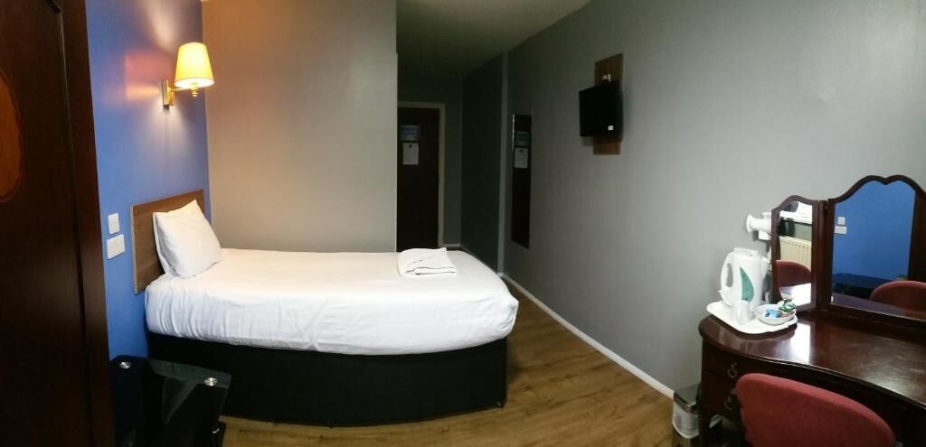 A hotel room with a bed, a desk with a chair, a mirror, a wall-mounted TV, and a small kitchenette area.