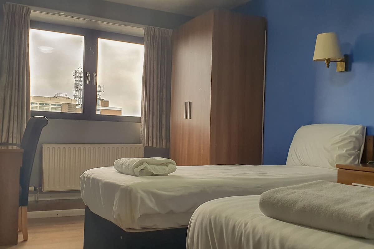 A hotel room with a bed, a nightstand, a lamp, a window with curtains, and a view of buildings outside.