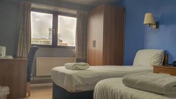 A hotel room with a bed, a nightstand, a lamp, a window with curtains, and a view of buildings outside.