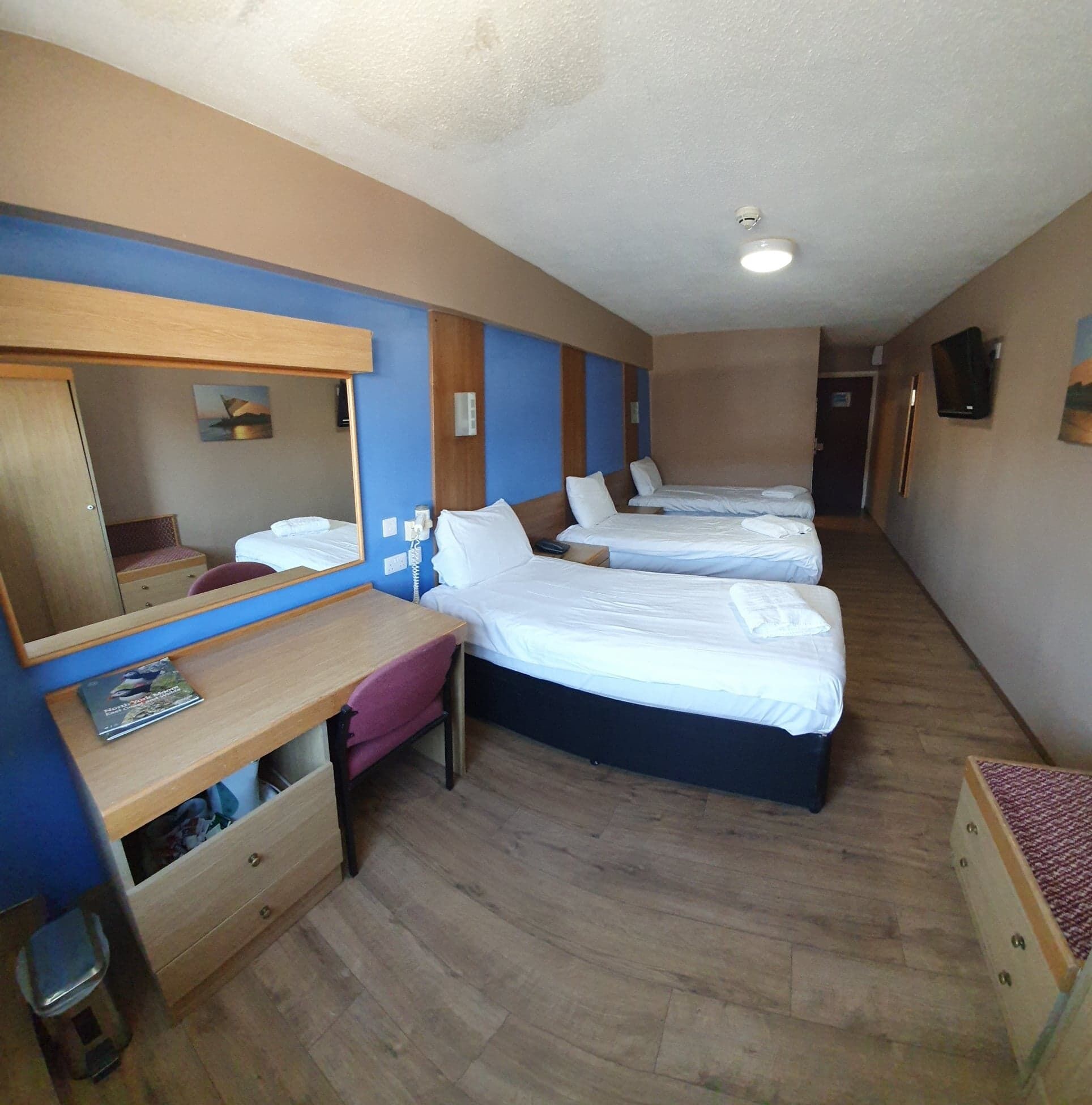 Quarto de hotel com duas camas, uma escrivaninha com cadeira, um espelho e uma lixeira pequena.