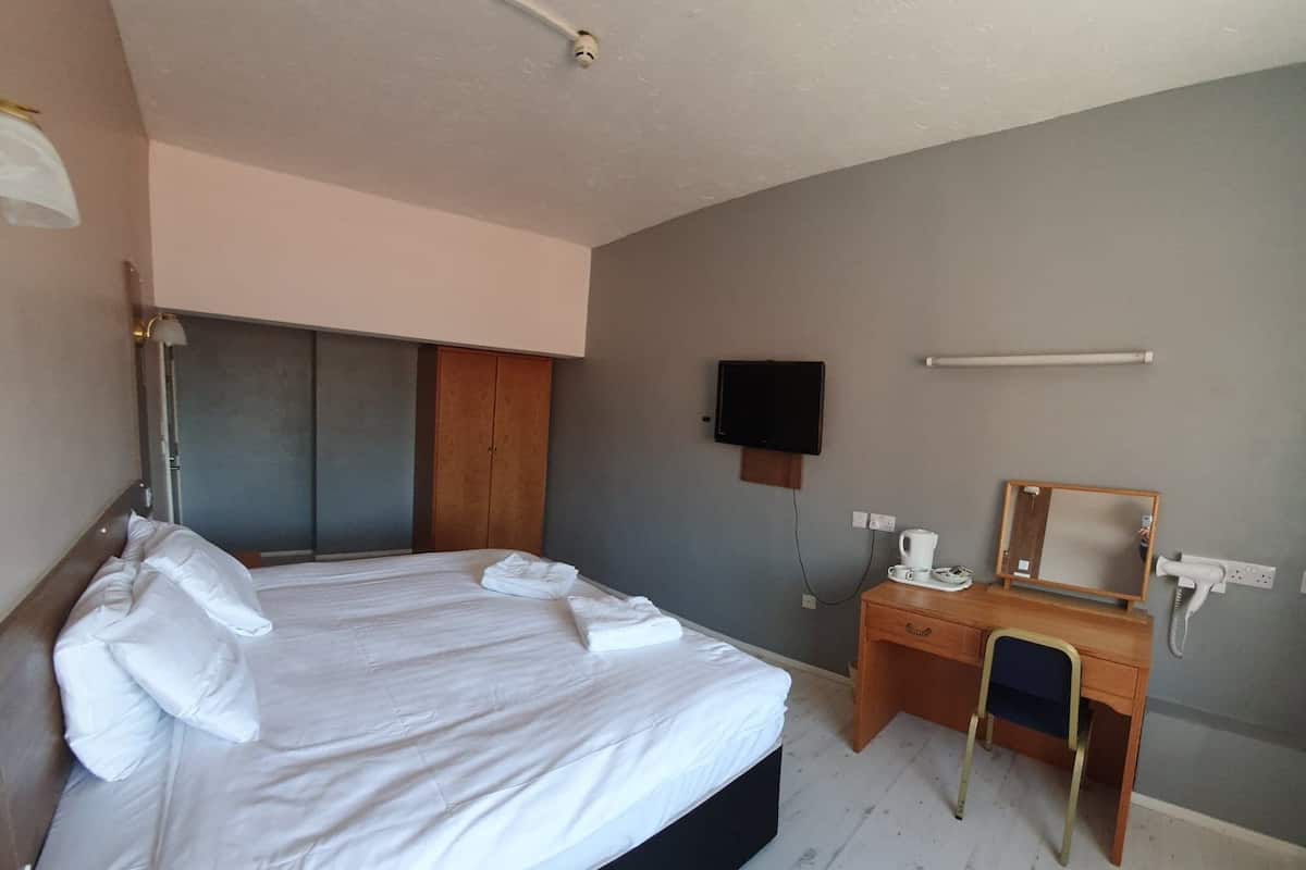 A neatly made bed with white linens and pillows, a wooden desk with a chair, a flat-screen TV mounted on the wall, and a hairdryer plugged in.