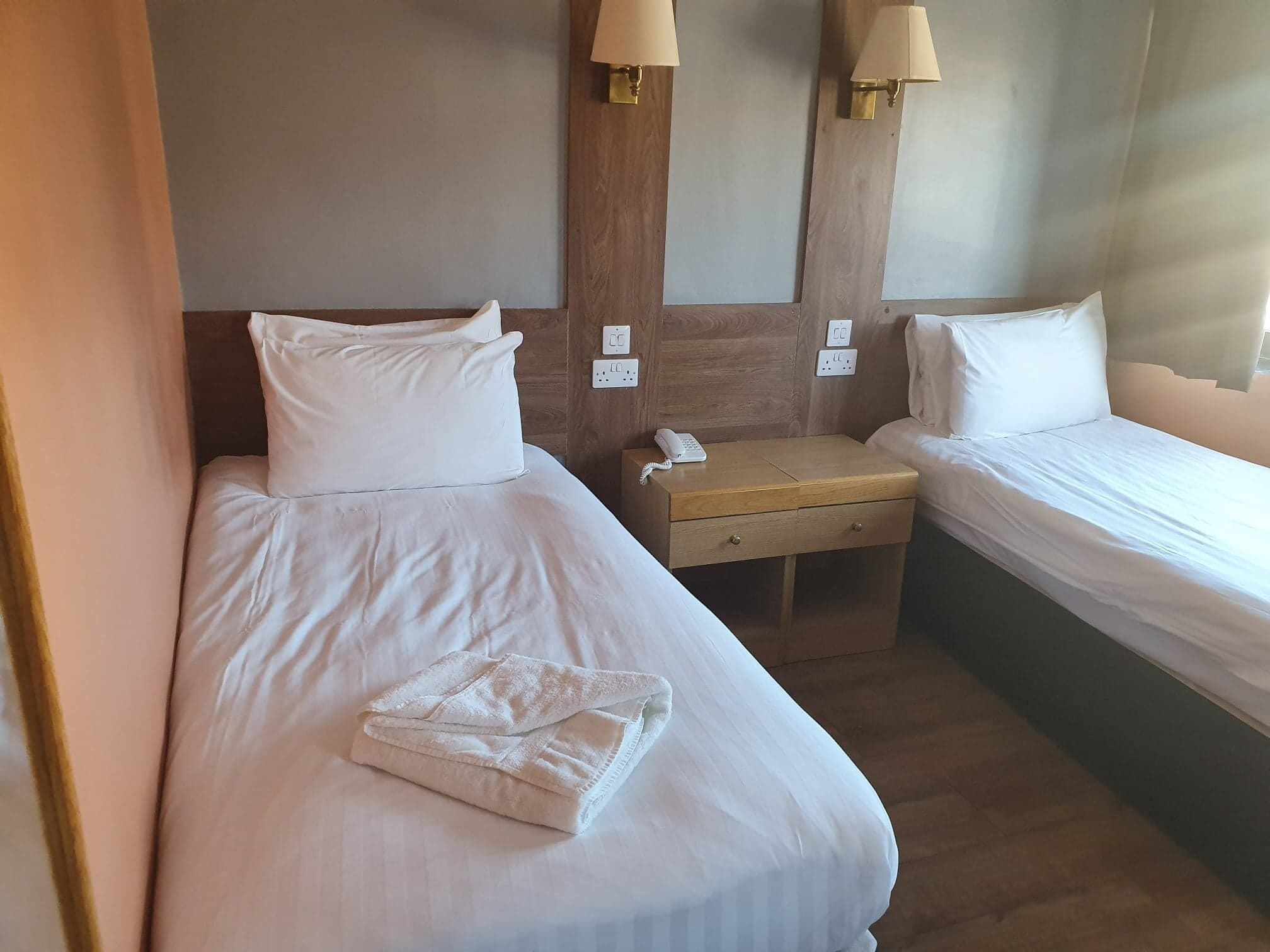 Une chambre d’hôtel avec deux lits simples, une table de chevet en bois avec un téléphone et des lampes fixées au mur.