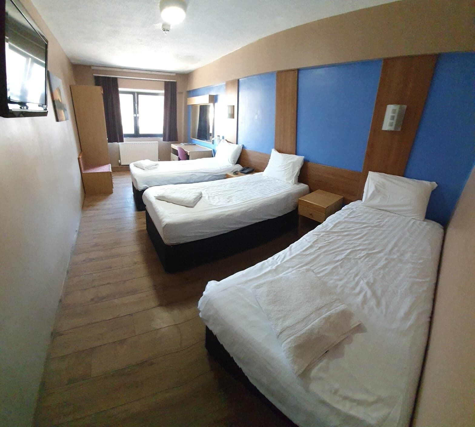 Habitación de hotel con tres camas, piso de madera, un escritorio con silla, televisión de pantalla plana y una ventana con cortinas.