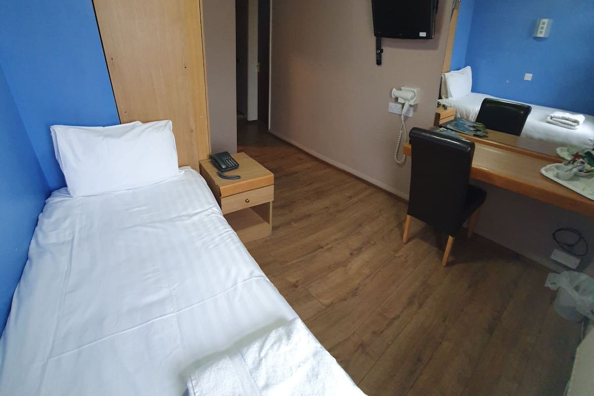A hotel room with a single bed, a desk with a computer, a chair, a telephone, and a TV mounted on the wall.