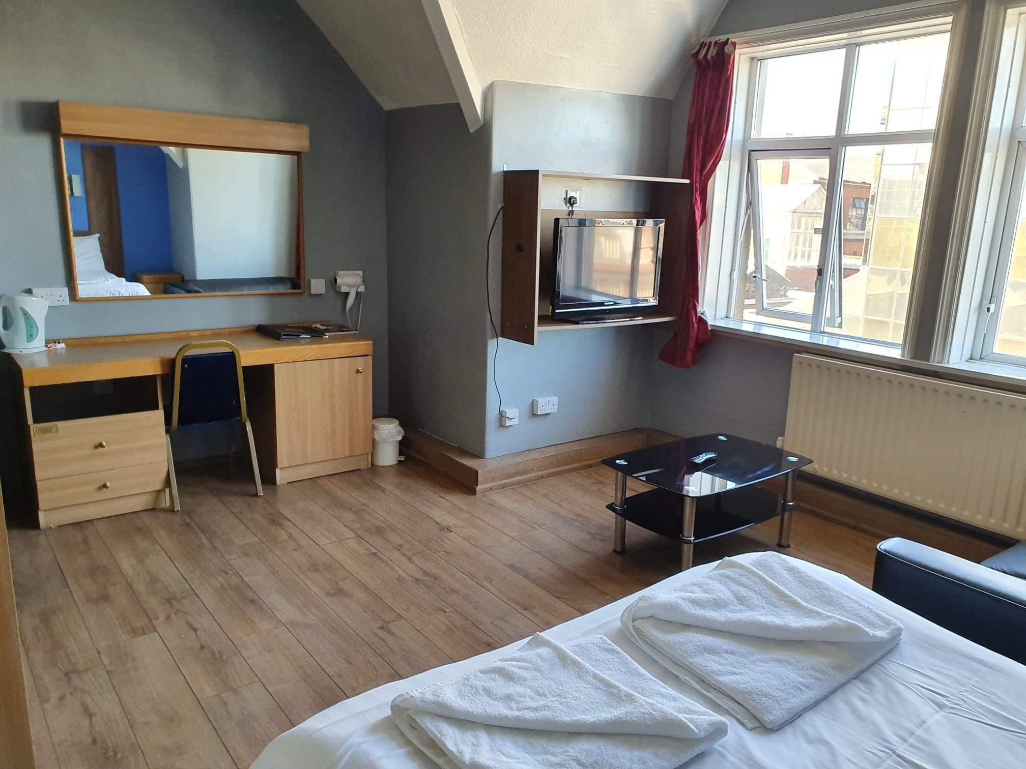 Una camera d'albergo con una scrivania in legno, una sedia, un letto con asciugamani bianchi, uno specchio, una TV e una finestra con tende.