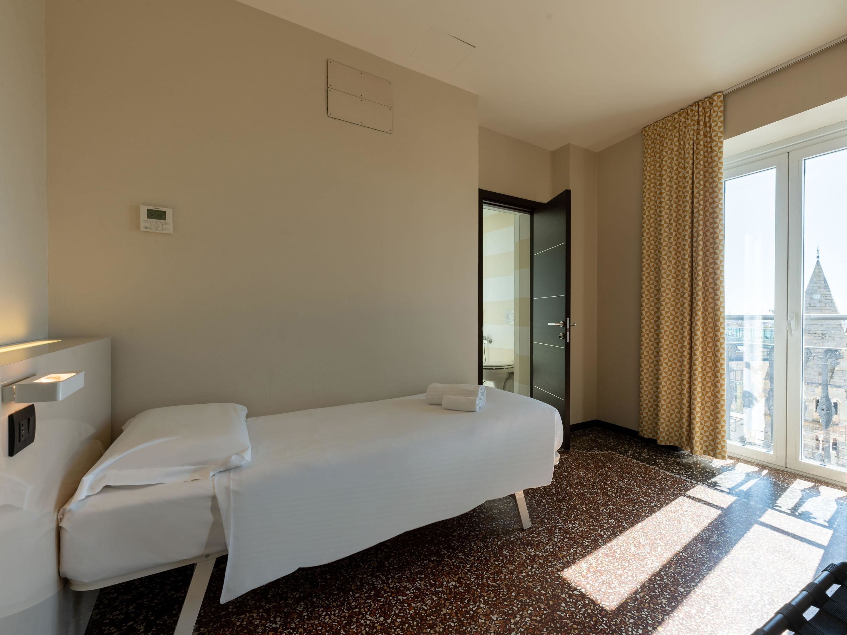 Ein Hotelzimmer mit einem Einzelbett, einem Nachttisch, einem Fenster mit Vorhängen und Blick auf ein Gebäude.