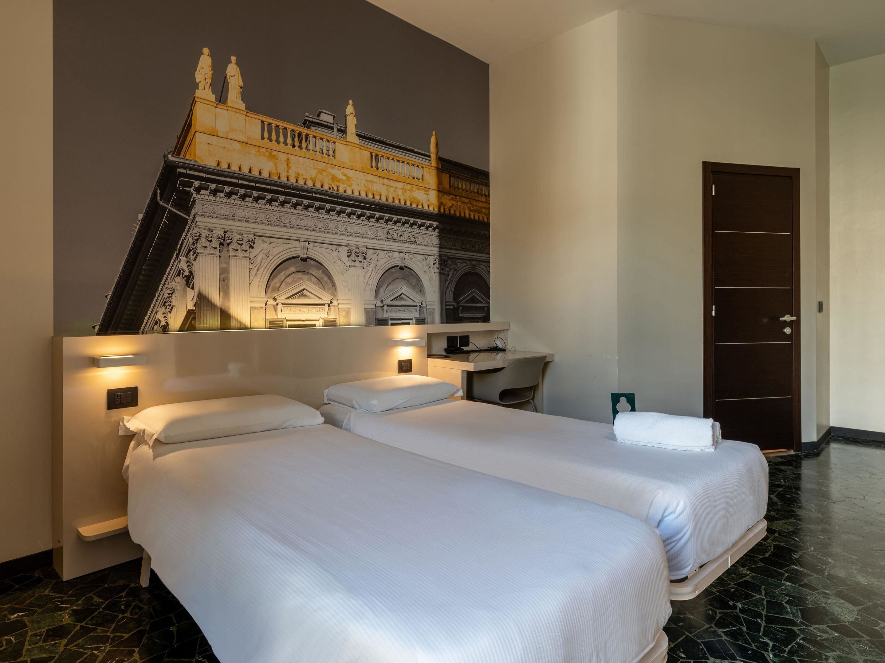 Een hotelkamer met twee bedden, een bureau en een groot muurschildering van een historisch gebouw.