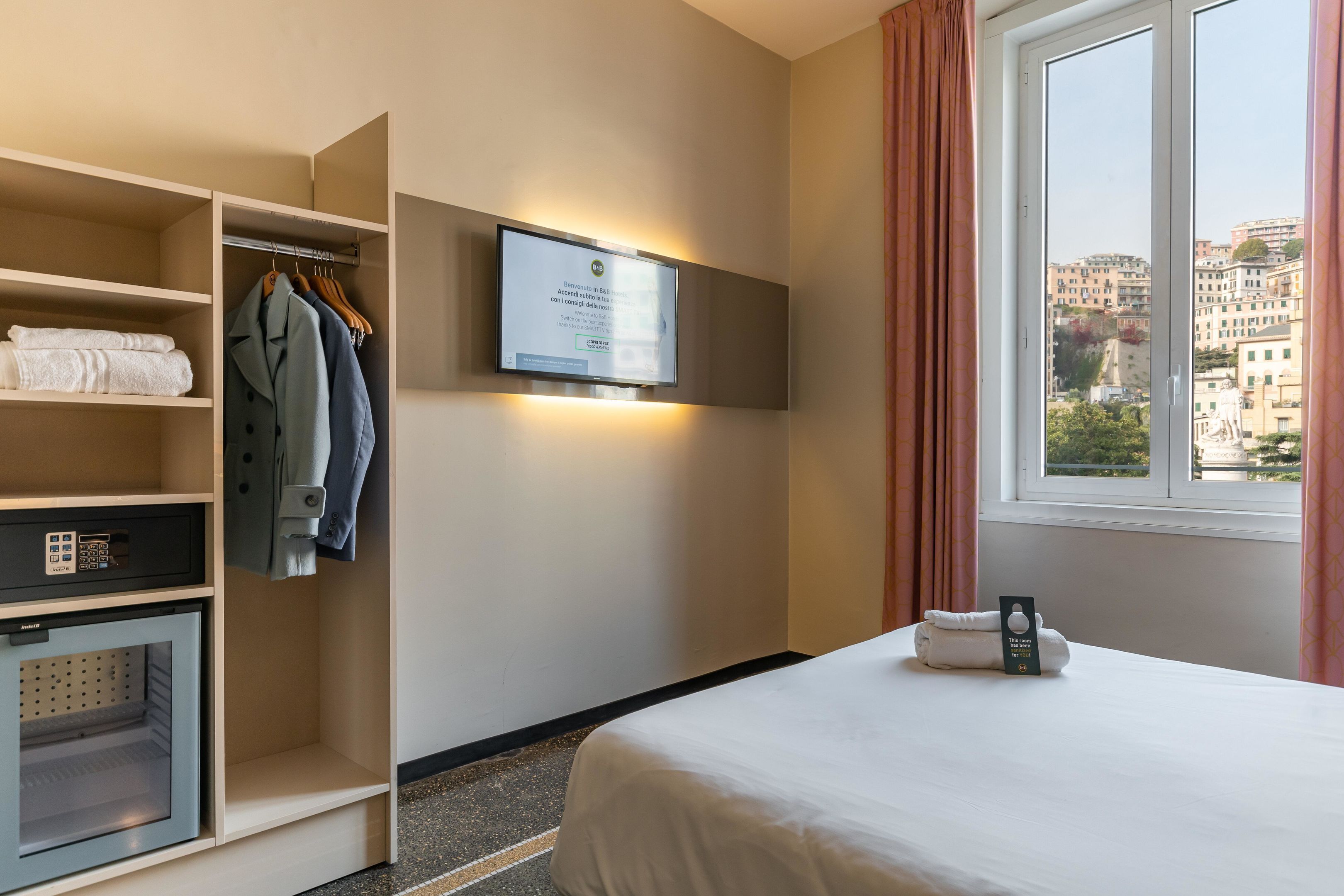 A hotel room with a bed, a wardrobe, a television mounted on the wall, and a window with curtains.