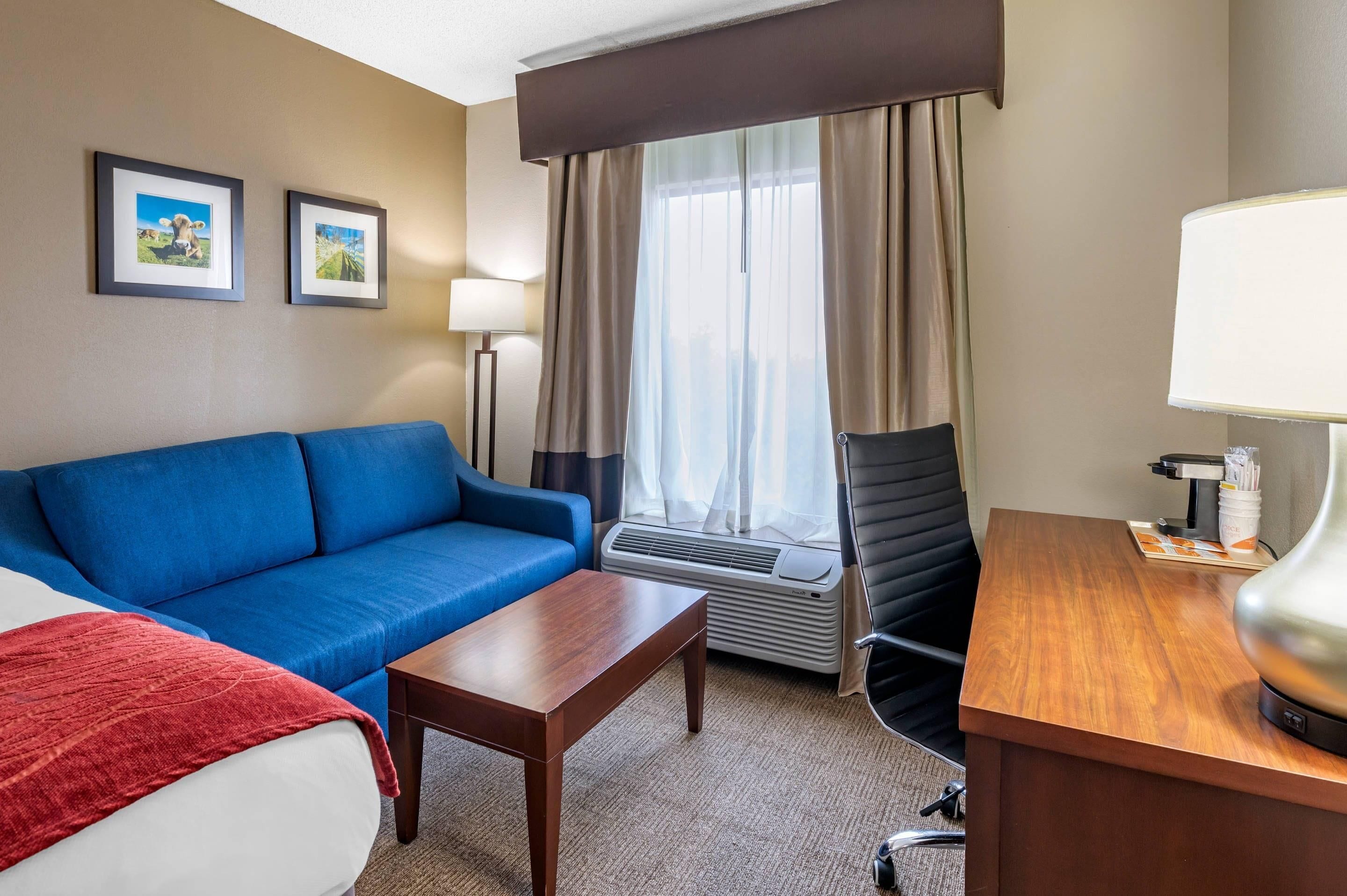 Habitación de hotel con sofá azul, escritorio de madera, una silla, cama, manta roja, lámpara, marco de fotos, una imagen, ventana con cortinas y unidad de aire acondicionado.