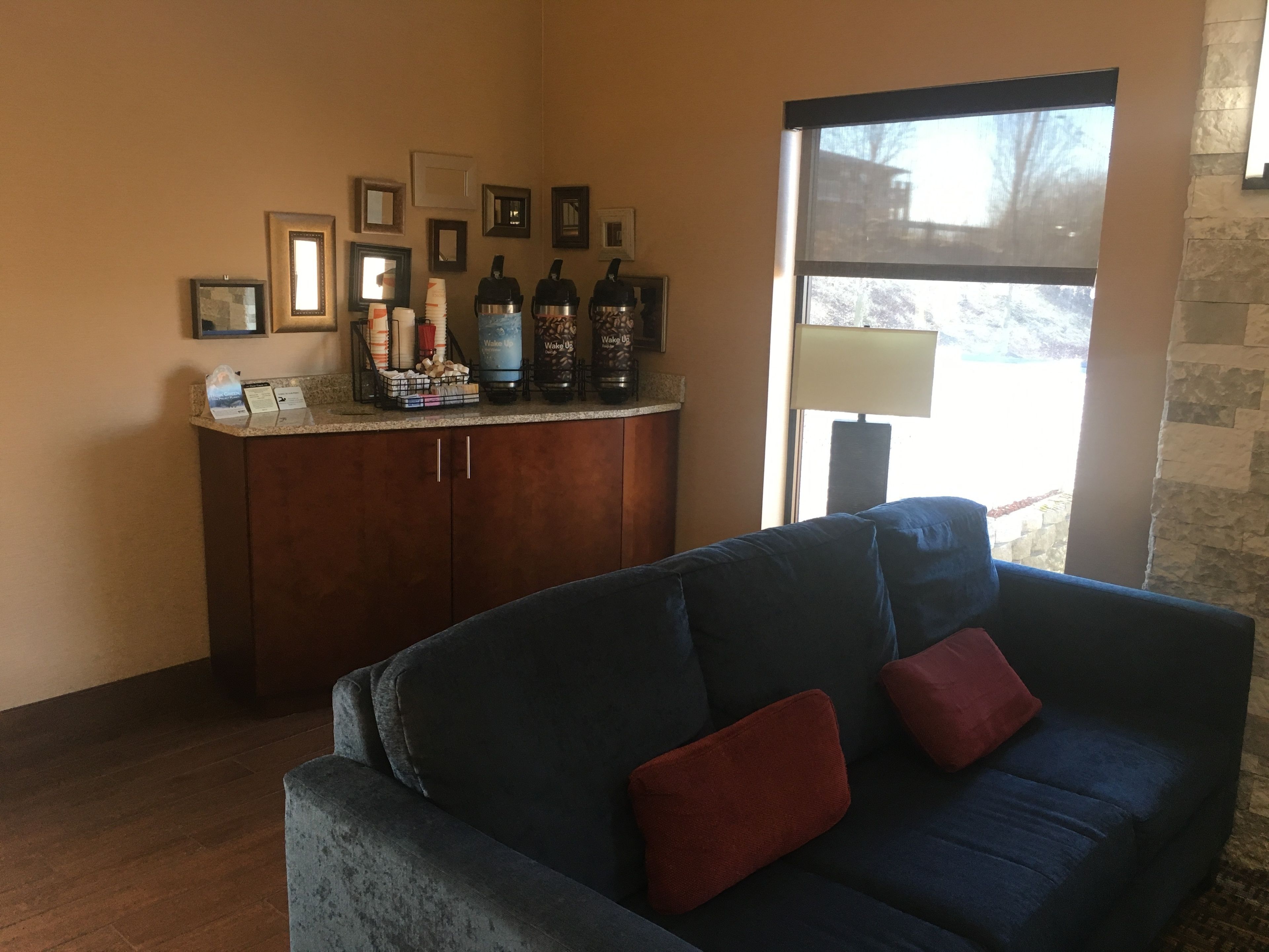 Una sala de estar con un sofá azul oscuro, un mueble de madera con cafeteras, cuadros enmarcados y una ventana con vistas a árboles.