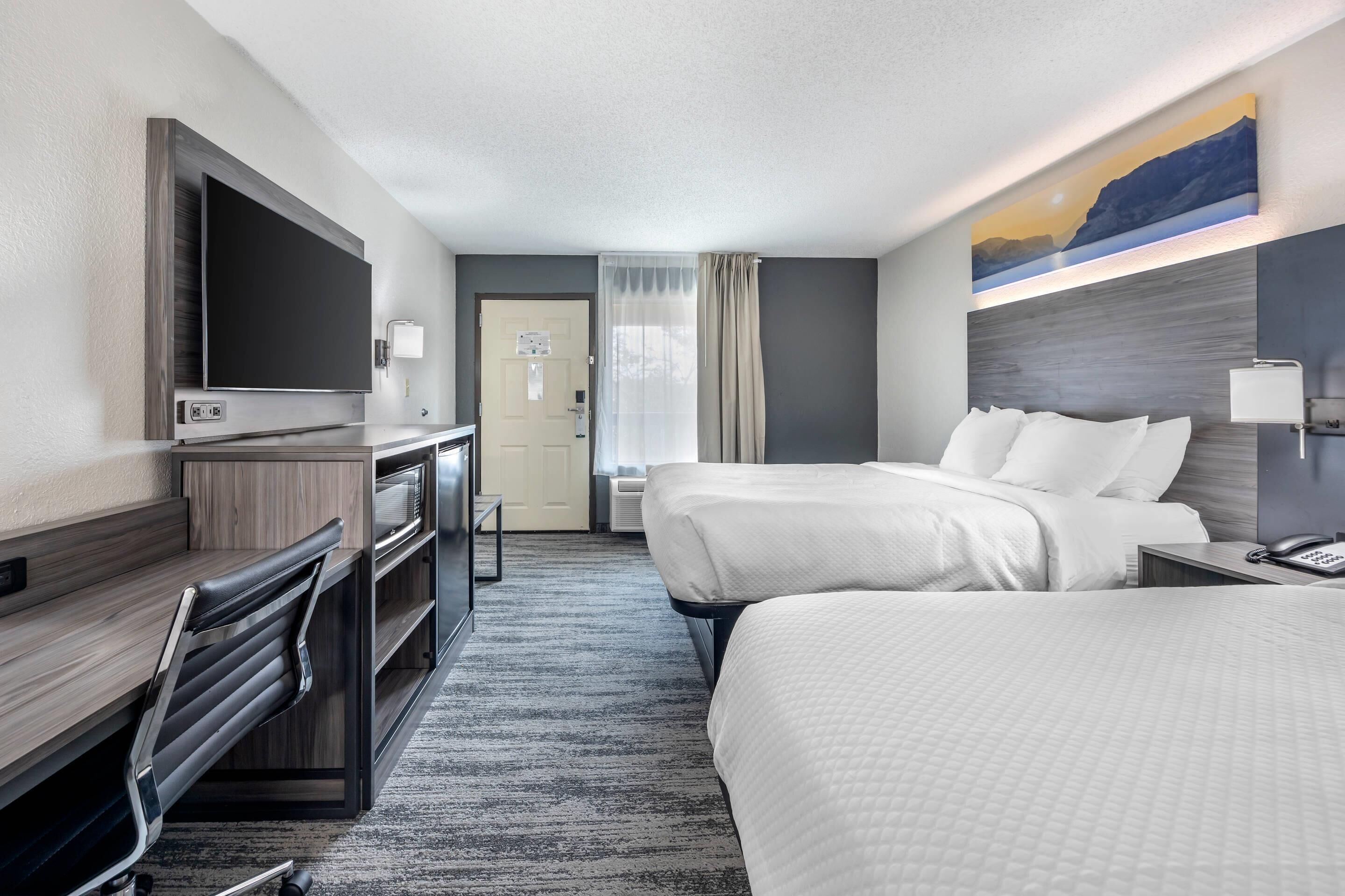 A hotel room with a large flat-screen TV, a desk with a chair, two beds, a painting on the wall, and a door with a curtain.