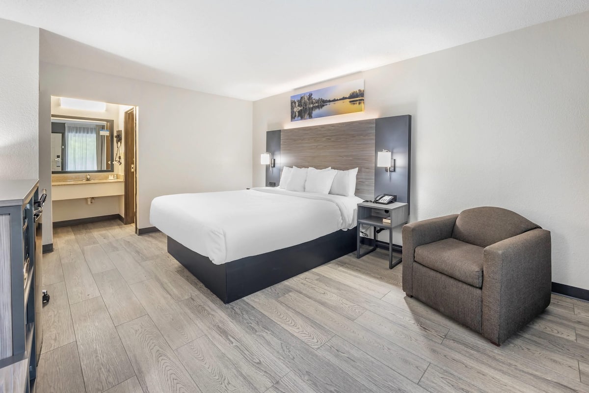 A modern hotel room with a large bed, a chair, a desk, and a nightstand.