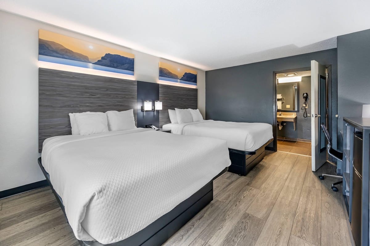 A hotel room with two beds, a large painting of a sunset, a wooden headboard, and a bathroom visible through an open door.