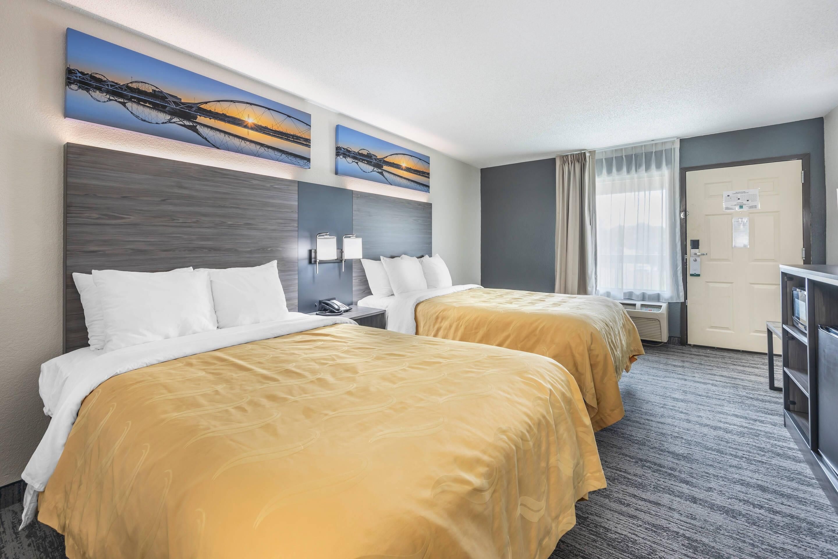 A hotel room with two beds, a desk, a chair, a TV, and artwork on the wall.