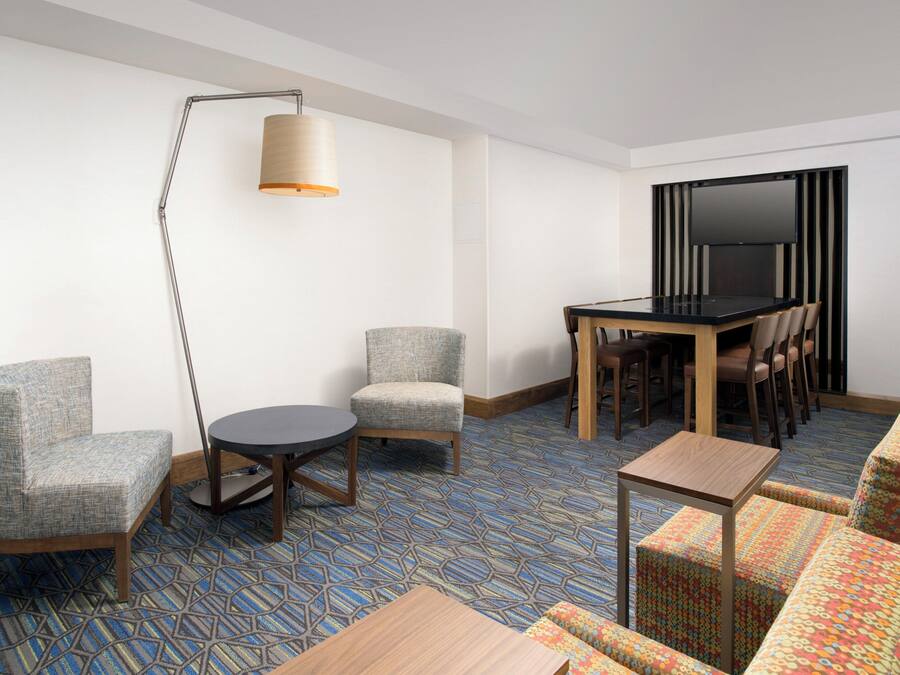 Quarto de hotel com um sofá, uma mesa redonda, uma mesa de jantar com cadeiras, uma TV na parede e um abajur.